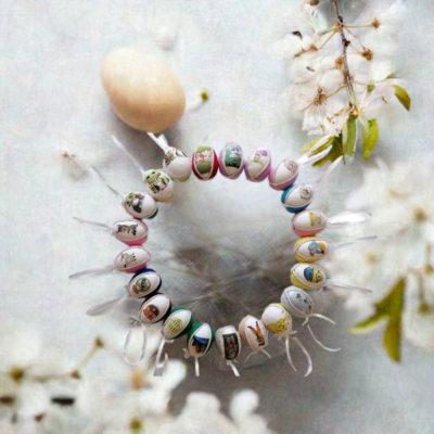 Kézzel készített húsvéti tojáskoszorú, amely színes, festett tojásokból áll, különböző vidám mintákkal és fehér szalagokkal díszítve. A háttérben tavaszi virágok láthatók, finom pasztell árnyalatokkal.