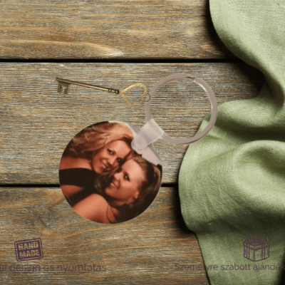Kerek, személyre szabott fényképes kulcstartó két mosolygó nő képével, egy fa felületen elhelyezve, mellette antik stílusú kulcs és zöld textil.